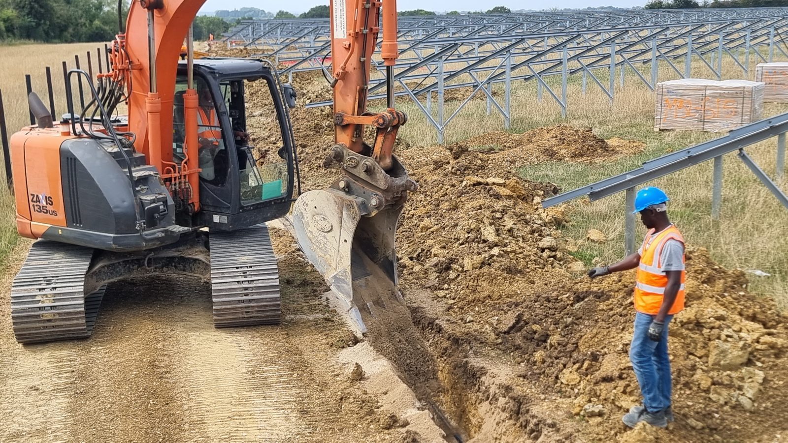 A tracked excavator digging a cable trench across a field at a renewable energy site.