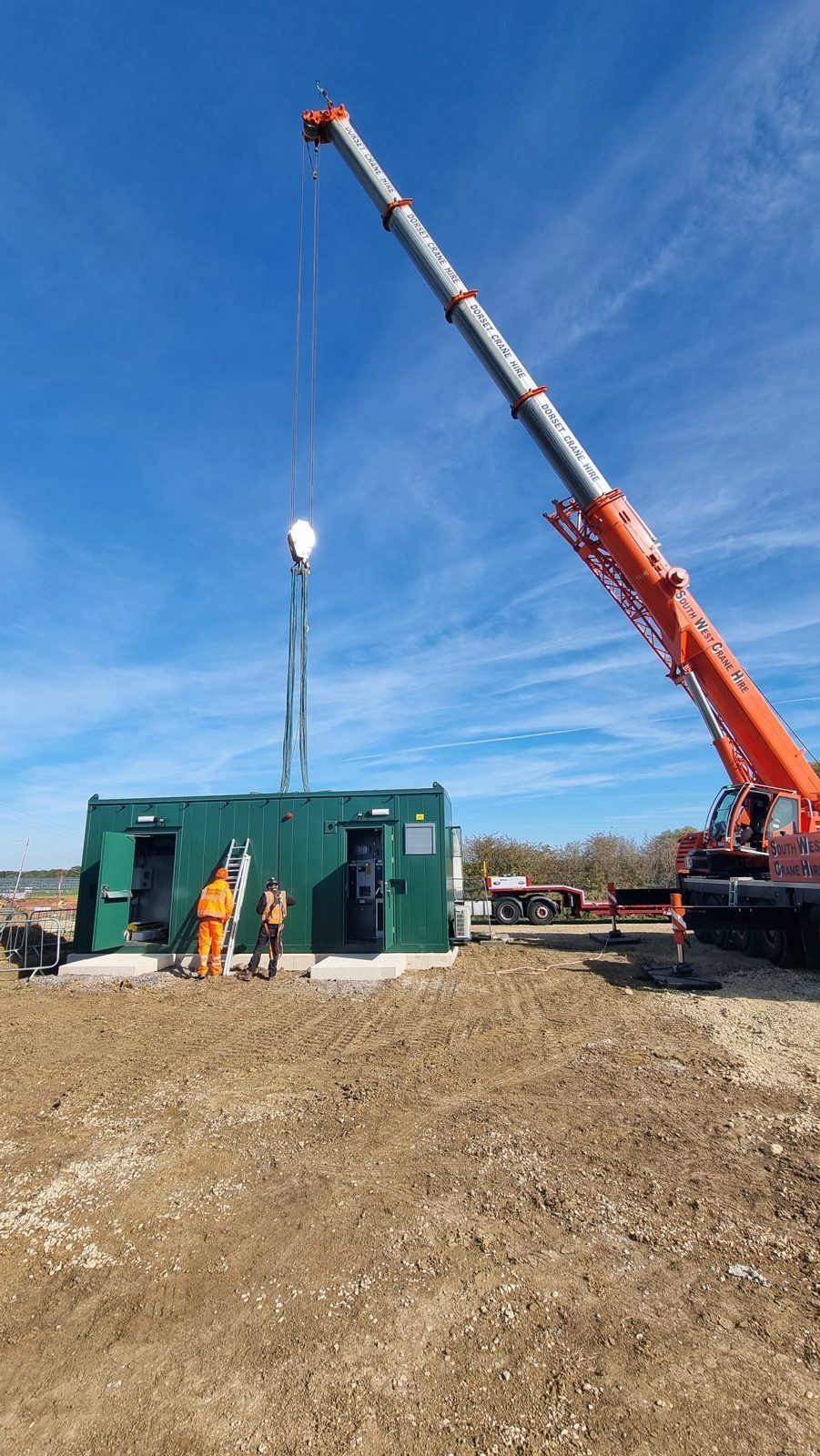 A mobile crane setting a combined switchgear and transformer unit onto its concrete base, with two HWBC operatives guiding the lift, at a renewable energy site in Cambridgeshire.