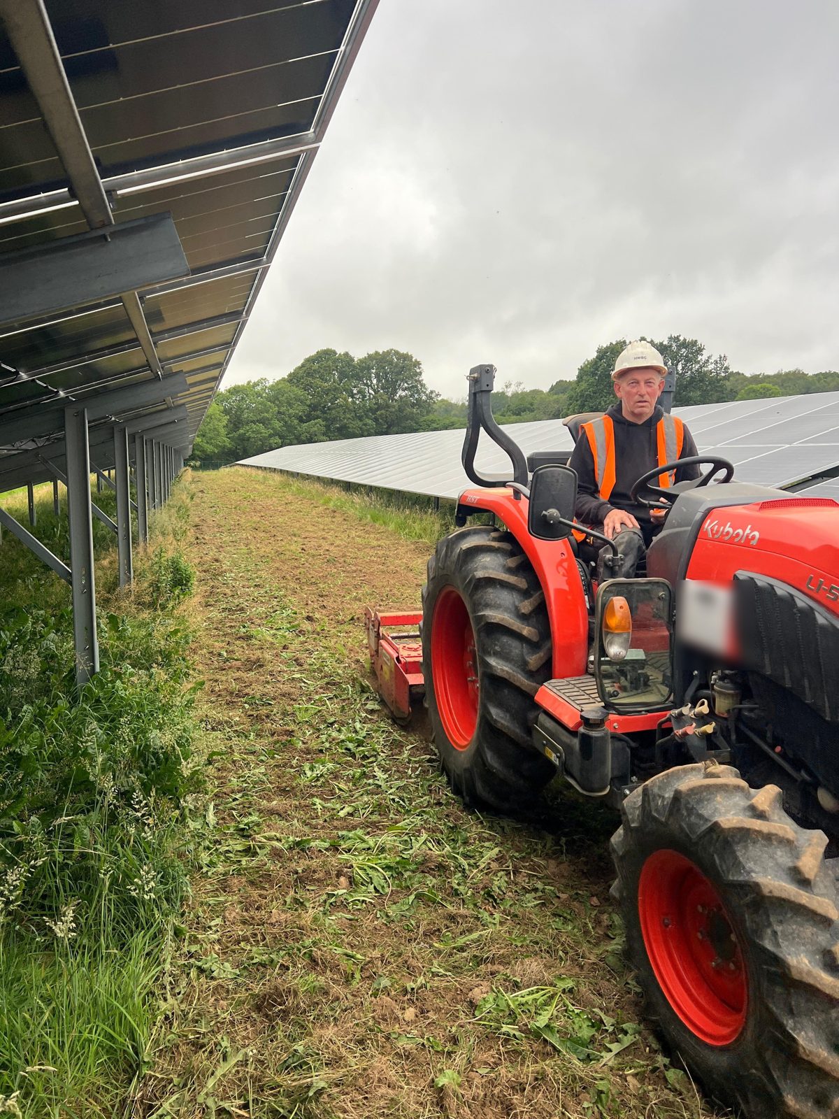 An HWBC operative driving a compact tractor and topper, managing the grass between solar panel rows in East Sussex.