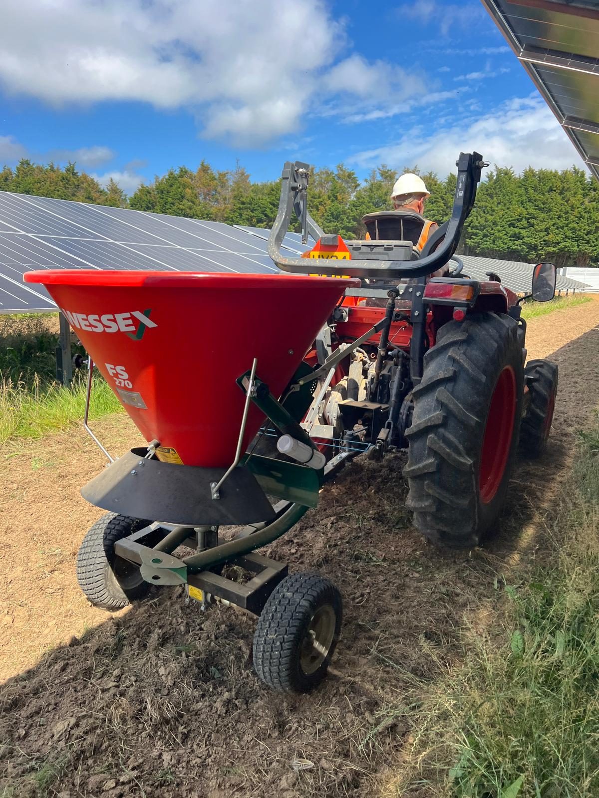 An HWBC operative driving a compact tractor and broadcast seeder between solar panel rows during reinstatement in East Sussex.
