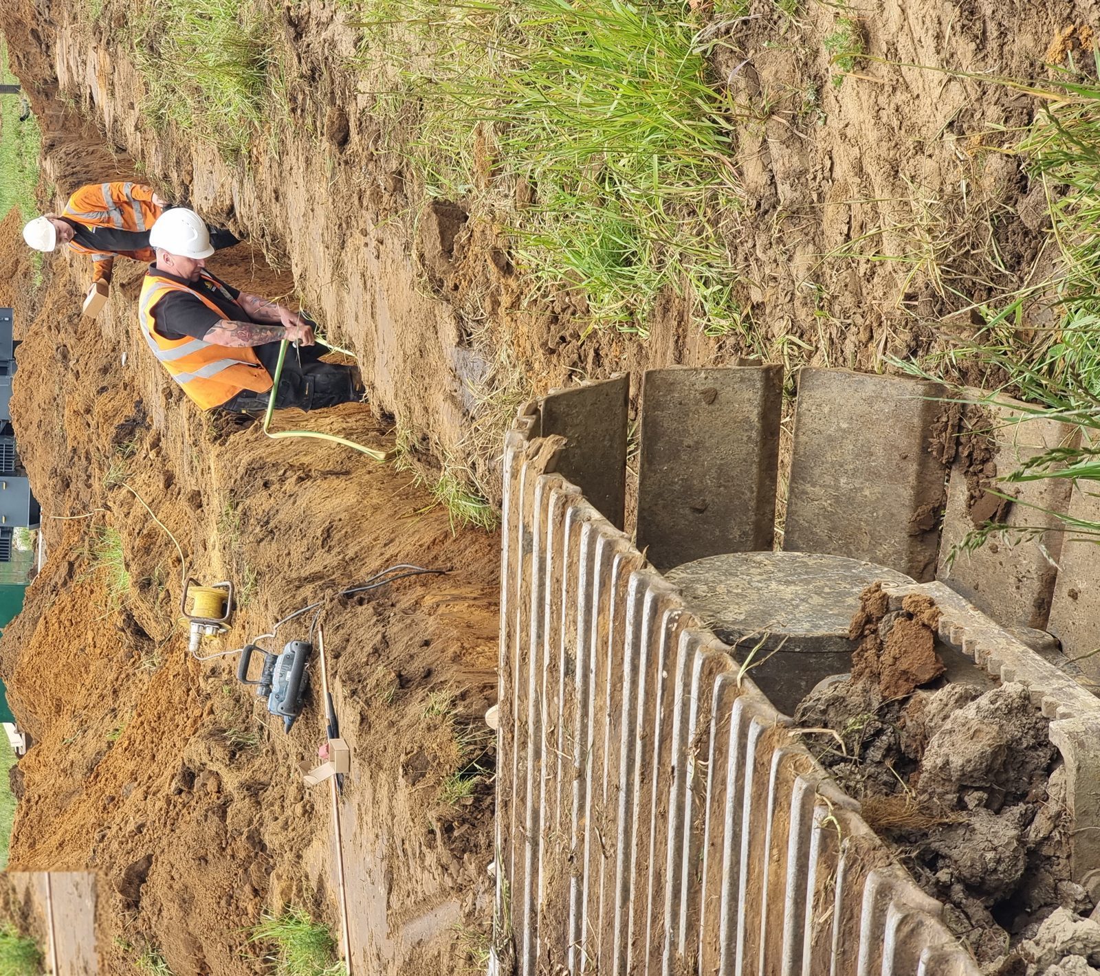 Two HWBC operatives installing an earthing system in a trench, with a transformer and green DNO connection cabinet behind, at a renewable energy site in West Northamptonshire.